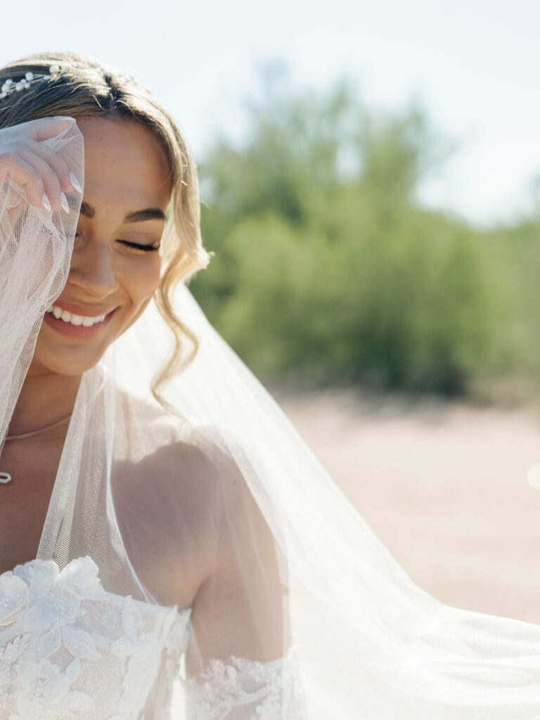 Bride smiling with her eyes closed, holding veil up next to her face.
