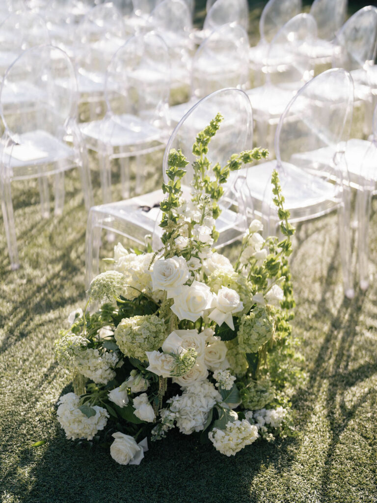 Ground floral arrangement of white and green flowers in wedding ceremony next to aisle chair.