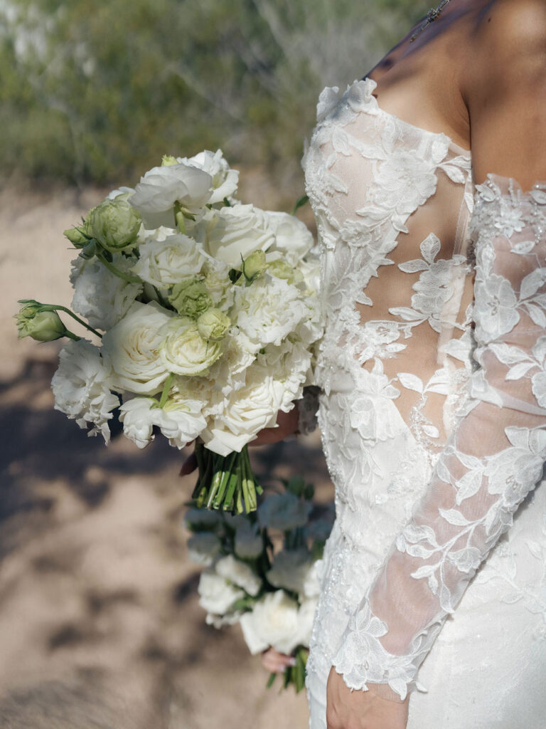 Bride in wedding gown with sheer upper-side holding bouquet of white roses and lisianthus.