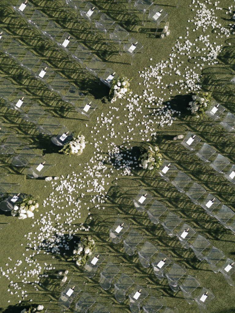 Overhear image of outdoor wedding ceremony with clear acrylic chair and white rose pedals down the center aisle.