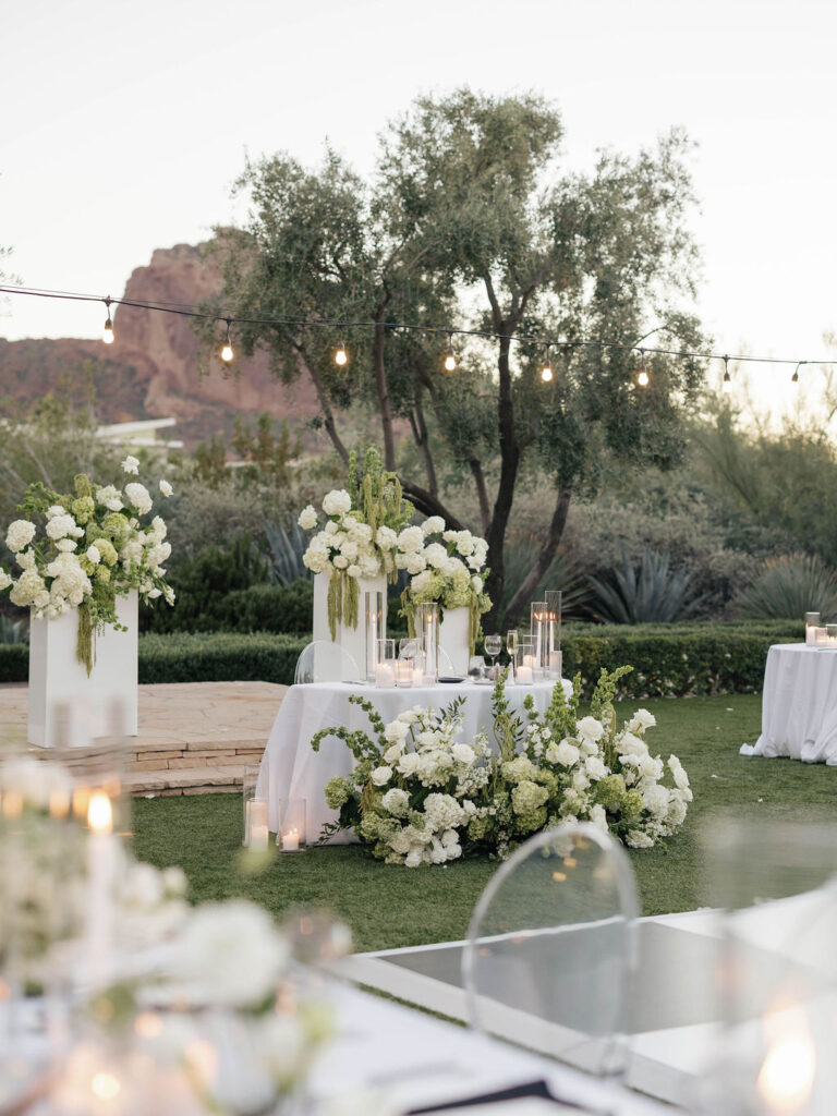 Sweetheart table design at outdoor reception with ground floral in front, candles on the table, and floral on columns behind.