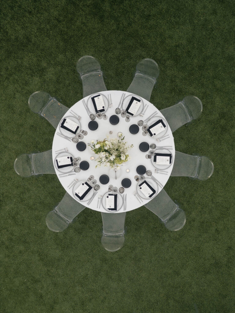 Overhead reception picture of round table with black and white place setting details, white floral centerpiece, and clear acrylic chairs.