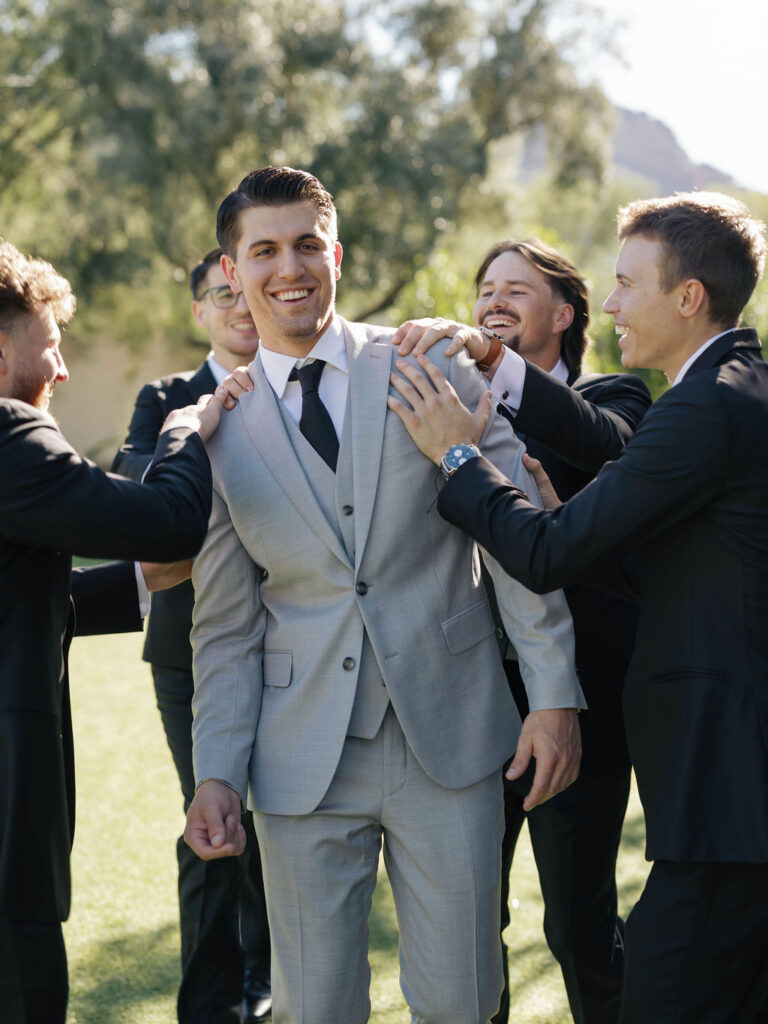 Groom in gray suit with groomsmen in black standing around him with their hands on his shoulders, all smiling.
