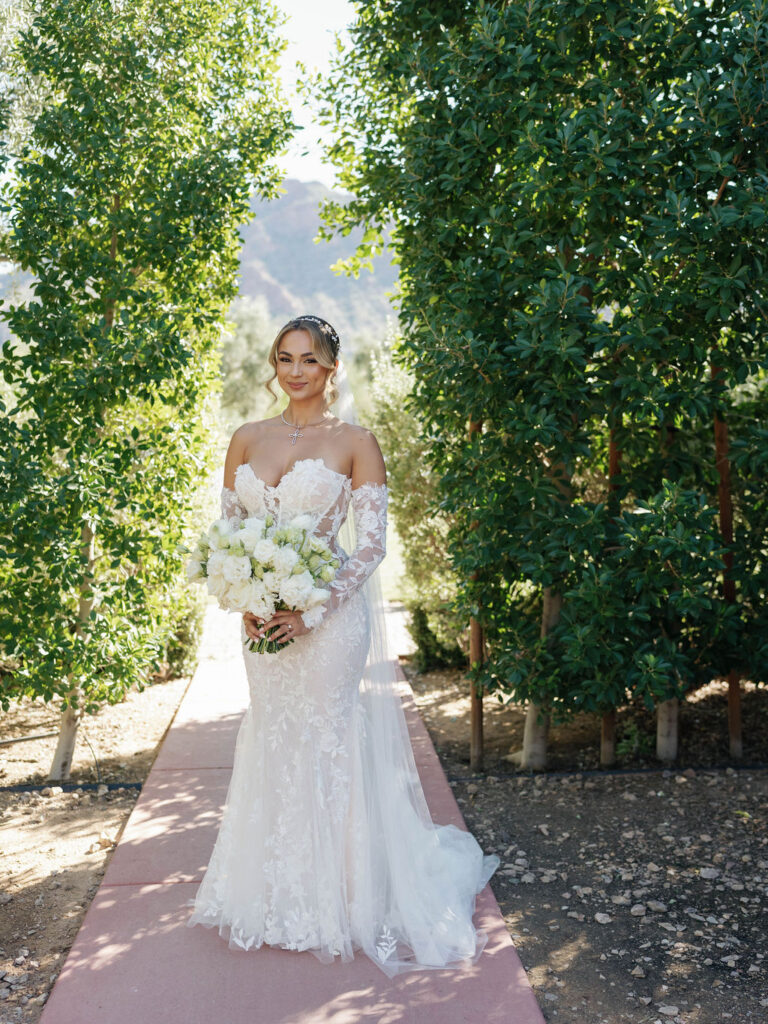 Bride standing in front of tall greenery on paved path, smiling, holding bouquet.