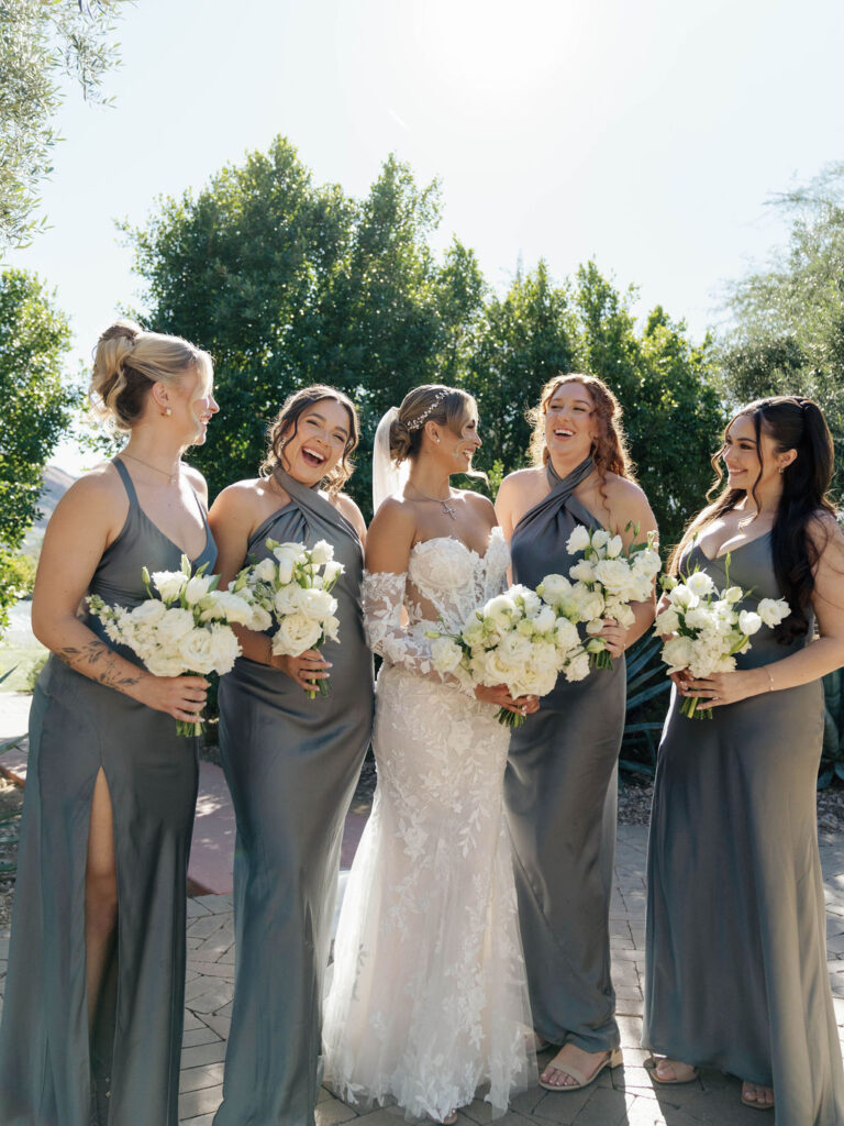 Bride with bridesmaids standing in a line, all smiling and holding bouquets of white flowers.