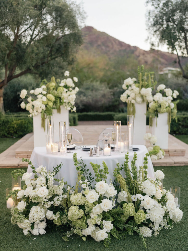 Sweetheart table design at outdoor reception with ground floral in front, candles on the table, and floral on columns behind.