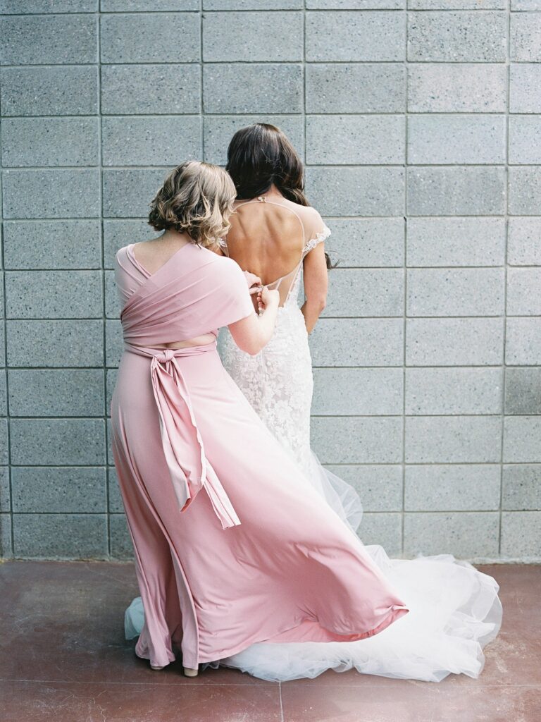 Bride facing away with wedding dress being buttoned by another woman in pink dress.