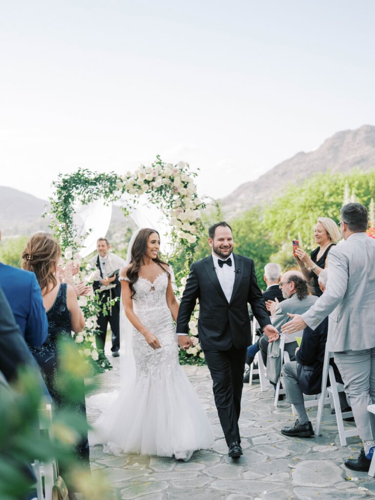 Bride and groom smiling, holding hands and walking down outdoor wedding ceremony aisle.