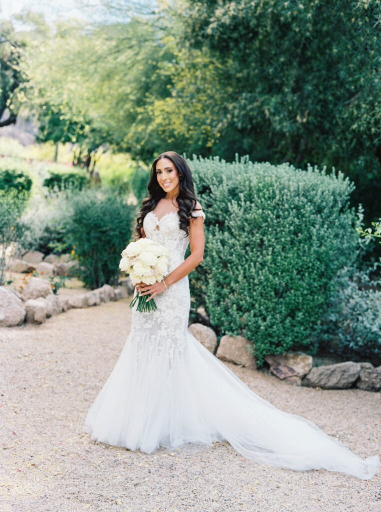 Bride standing in desert landscape holding bouquet of white and blush roses, smiling.