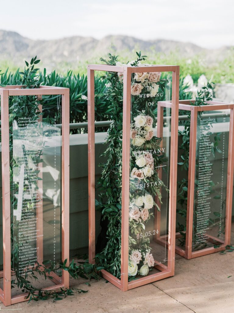 Custom wedding welcome and escort signs of clear acrylic and floral with greenery suspended in center with pink metal frames.
