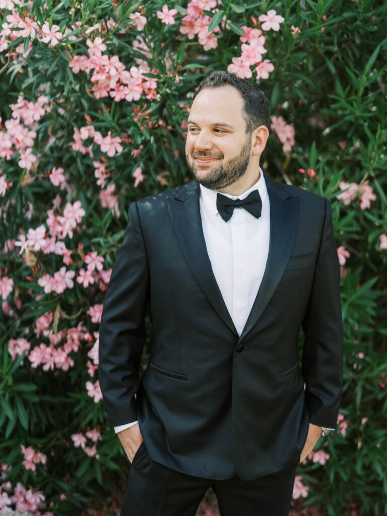 Groom smiling and looking off to side, standing with hands in pockets of black tux in front of large flowering plant.