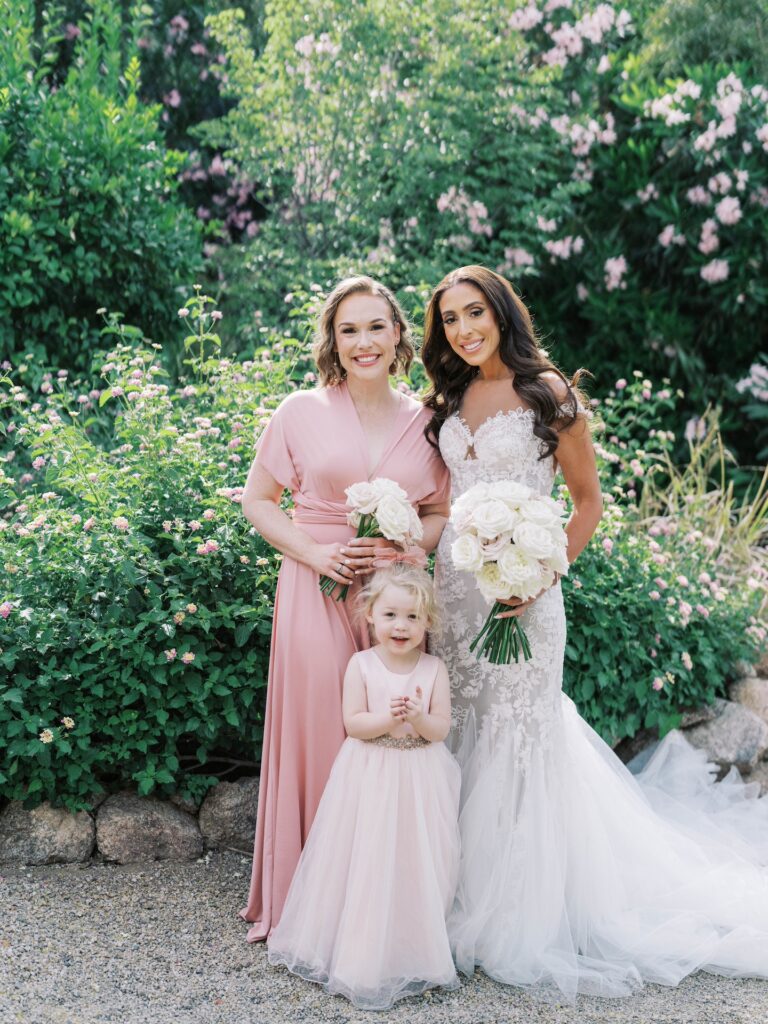 Bride standing with woman both holding bouquets, and young girl in blush pink dresses, all smiling.