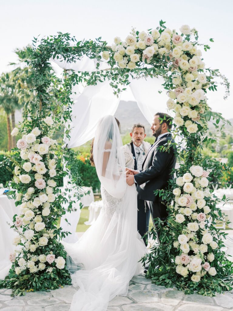 Bride and groom holding hands and standing under wedding ceremony chuppah with officiant.