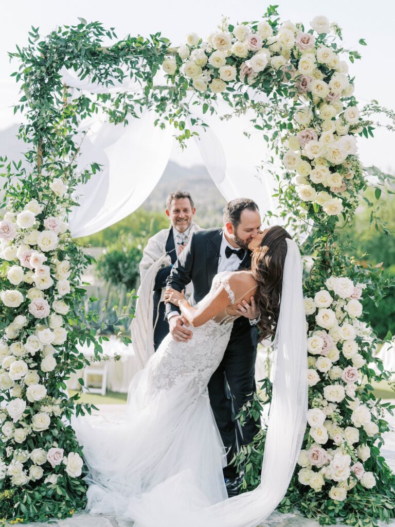 Groom dipping bride and kissing her under chuppah with flowers and greenery.