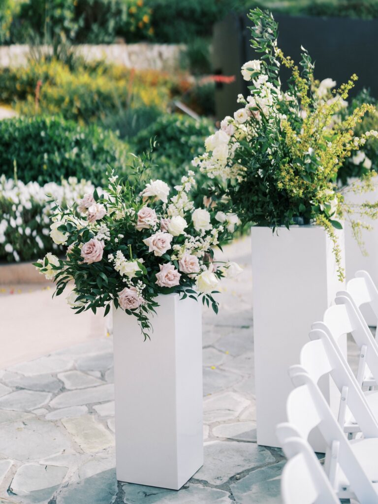 Back of aisle wedding ceremony floral arrangements of blush and white flowers and greenery on white acrylic columns.