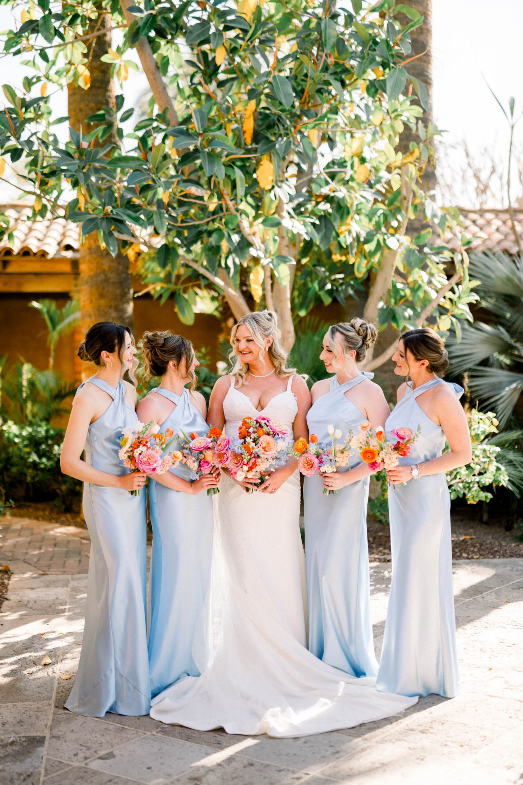 Bride standing with bridesmaids in baby blue gowns all holding colorful floral bouquets.