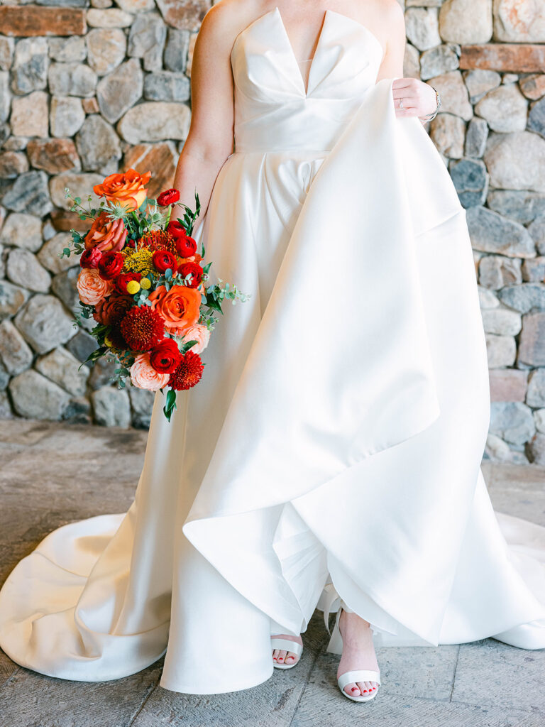 Bride in gown holding bouquet of red, pink, orange, and peach flowers.
