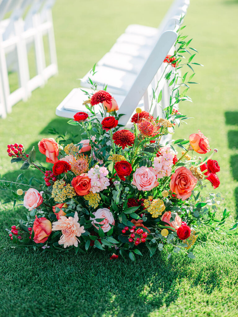 Wedding ceremony ground floral placed in ceremony aisle next to chair in flowers of pink, peach, red, and orange with greenery.