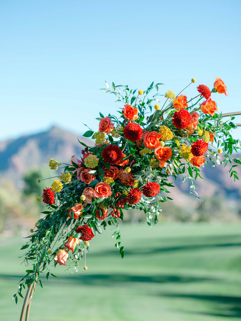 Flowers of red, peach, yellow, and orange with greenery installed on wedding ceremony circle arch.