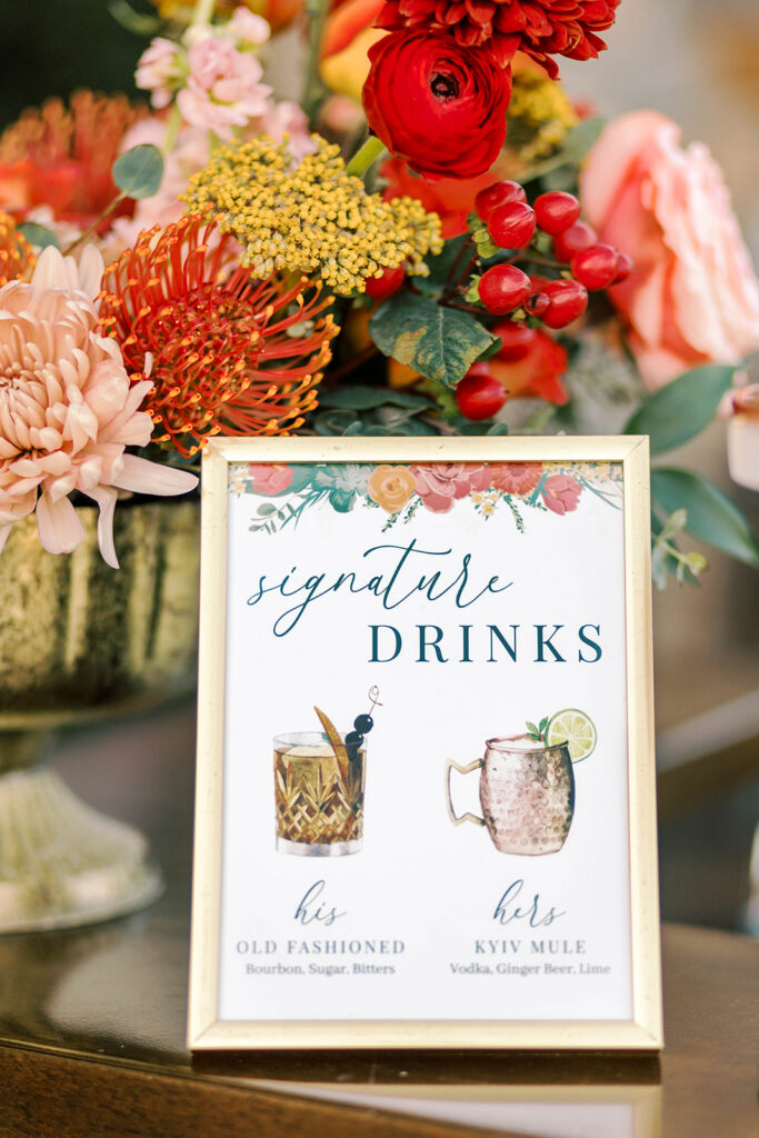 Custom cocktail sign at wedding bar with floral arrangement behind it.
