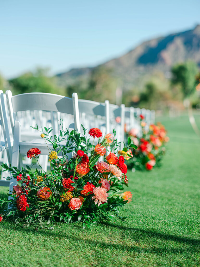 Wedding ceremony ground floral placed in ceremony aisle next to chair in flowers of pink, peach, red, and orange with greenery.