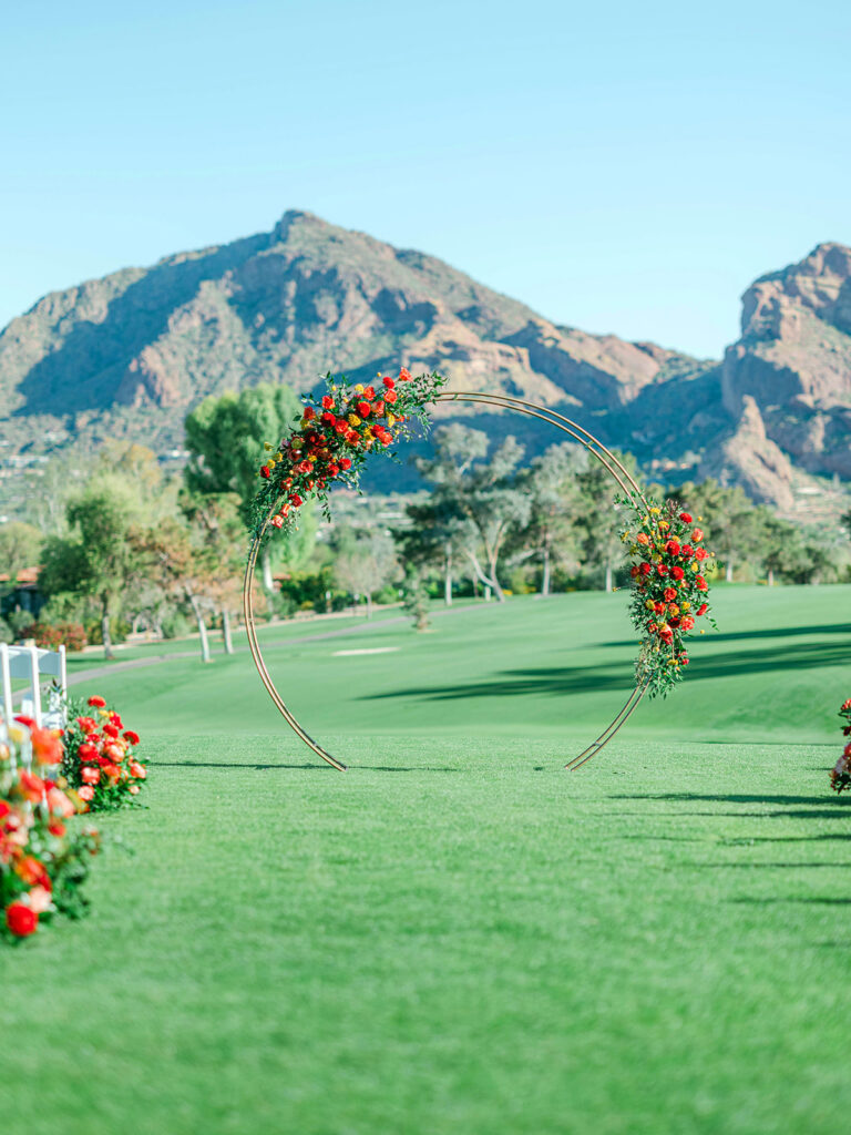 Outdoor wedding ceremony gold circle arch in front altar space with two asymmetrical floral arrangements installed on it. Mountains in background.