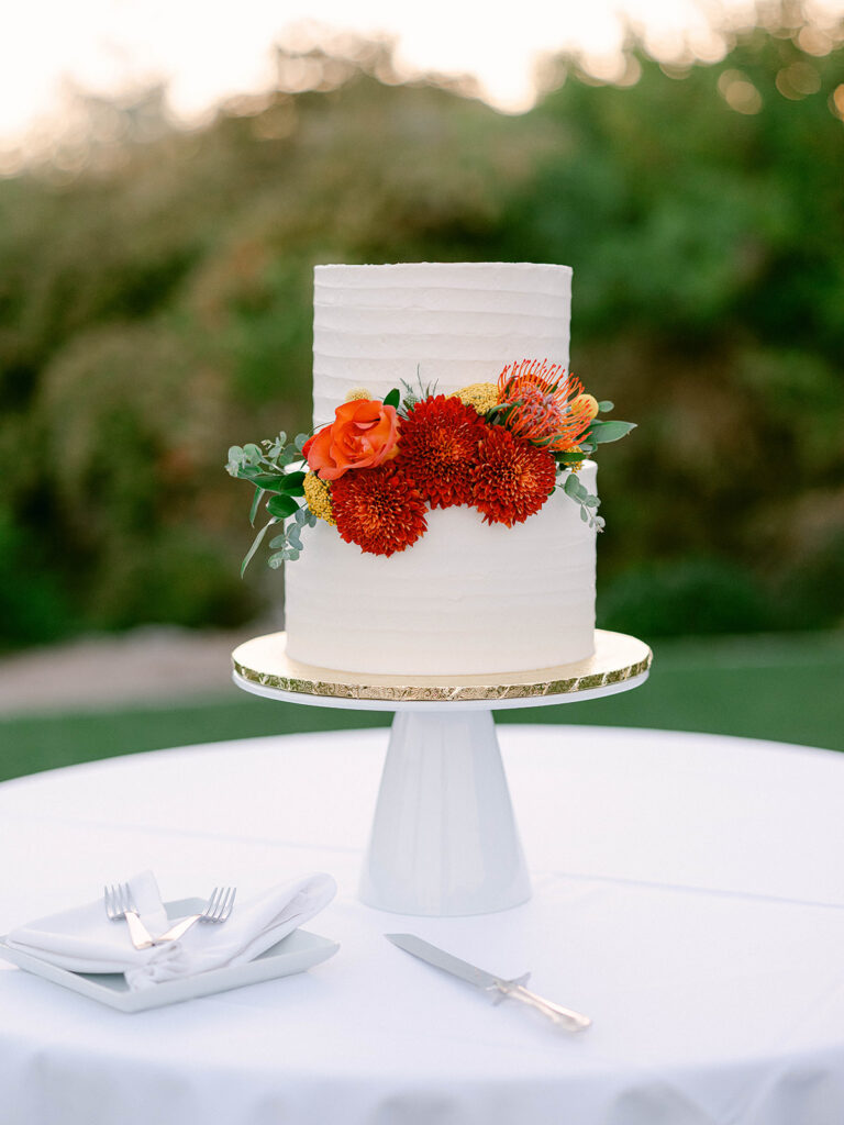 White two tiered wedding cake on white stand with red, peach, and yellow colored flowers placed together on top of bottom layer.